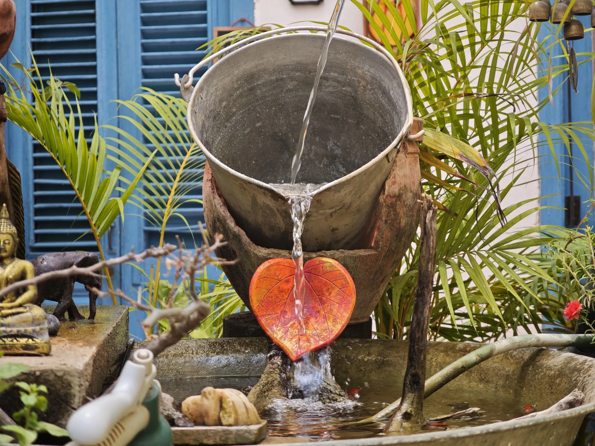 Fuente con un cubo de metal viejo que vierte agua sobre una hoja naranja en forma de corazón, entre palmas